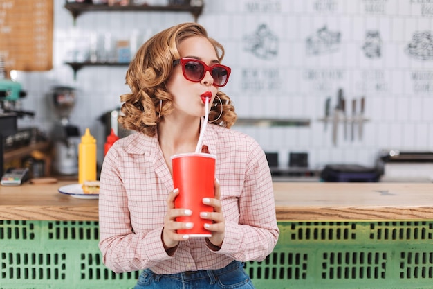 Bella signora in occhiali da sole e camicia seduta al bancone del bar e bere acqua gassata mentre guarda da parte nella caffetteria