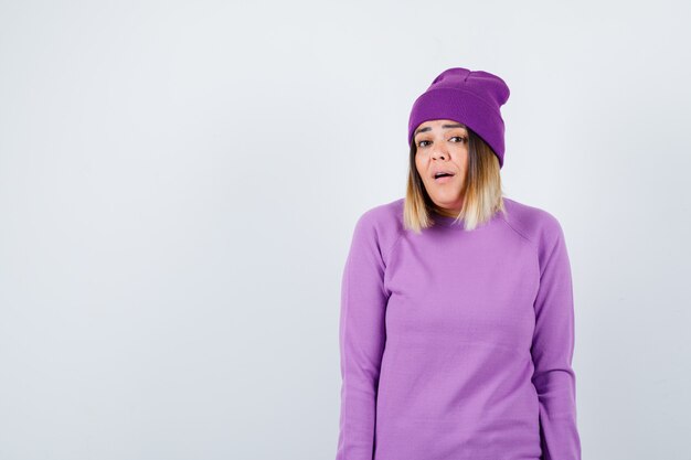 Beautiful lady in sweater, beanie posing while standing, opening mouth and looking surprised , front view.