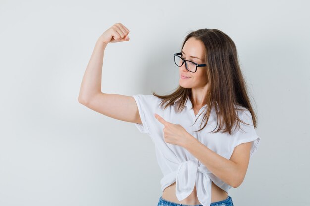 Beautiful lady in white blouse,glasses pointing at arms muscles and looking confident , front view.