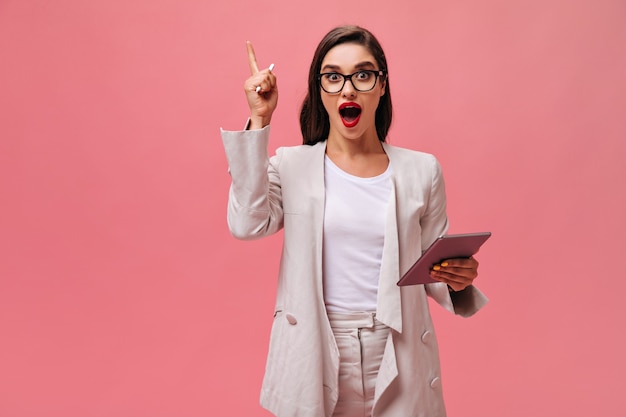 Beautiful lady in white stylish suit and eyeglasses has cool idea and poses with computer tablet on isolated pink background.