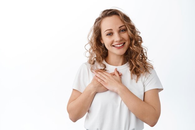 Beautiful lady with blond curly hair holding hands on heart and looking grateful say thank you express gratitude standing over white background