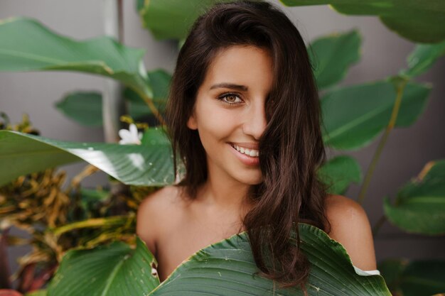 Beautiful lady with curly hair and green eyes smiles sweetly Woman with long eyelashes posing against backdrop of tropical plant