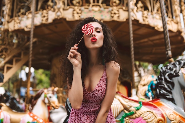 Beautiful lady with dark curly hair in dress standing and covering her eye with lolly pop candy while dreamily looking in camera with carousel on background