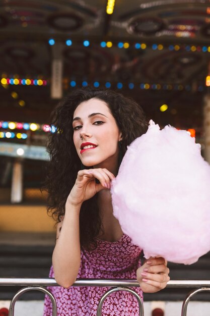 Beautiful lady with dark curly hair in dress standing with cotton candy in hand and dreamily looking in camera while spending time in amusement park