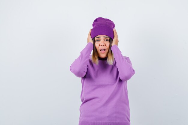 Beautiful lady with hands on head in sweater, beanie and looking horrified. front view.