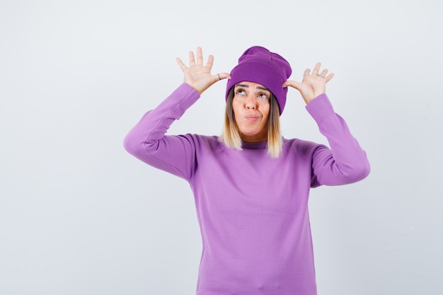 Foto gratuita bella signora con le mani vicino alla testa come orecchie in maglione, berretto e dall'aspetto divertente, vista frontale.