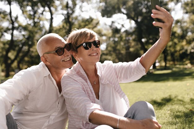 Beautiful lady with short hair in sunglasses, pink blouse and jeans sitting on grass and making photo with grey haired man in white outfit on park.