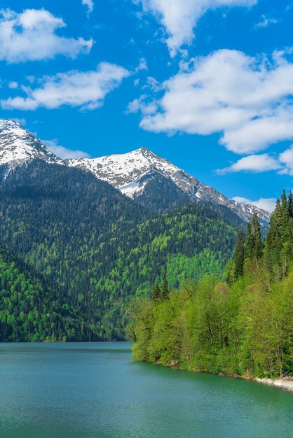 Красивое озеро Рица в горах Кавказа. Зеленые холмы горы, голубое небо с облаками. Весенний пейзаж.