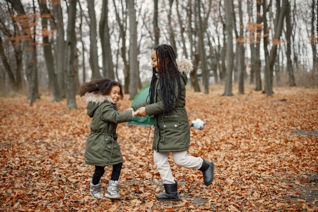 Beautiful little black girls standing near tent in the forest Two little sisters dancing in autumn forest Black girls wearing khaki coats