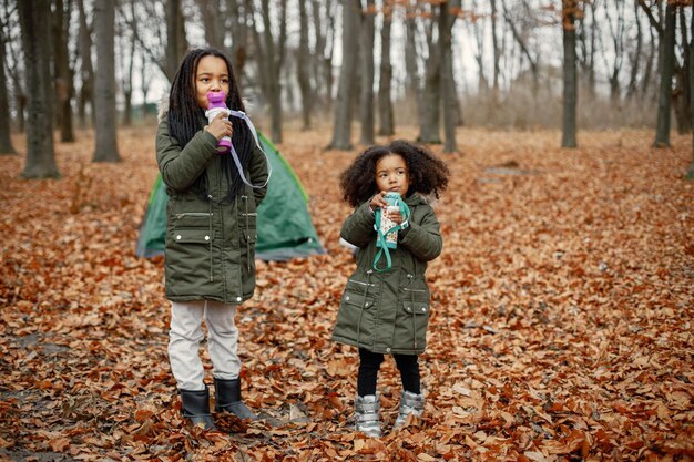 森のテントの近くに立っている美しい小さな黒人の女の子秋の森の魔法瓶からお茶を飲む2人の妹カーキ色のコートを着た黒人の女の子