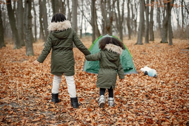森のテントの近くに立っている美しい小さな黒人の女の子秋の森で遊んでいる2人の妹カーキ色のコートを着ている黒人の女の子