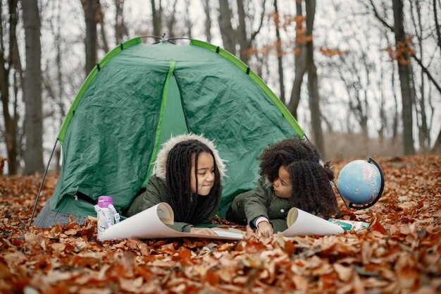 Beautiful little black girls in tent camping in the forest Two little sisters lying in a tent in autumn forest and looking on a map Black girls wearing khaki coats