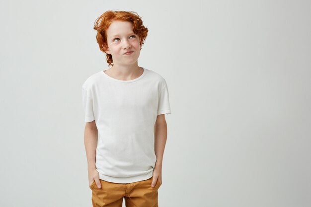 Beautiful little ginger kid in white t shirt and yellow jeans holding hands in pockets, looking aside with funny expression planning something bad.