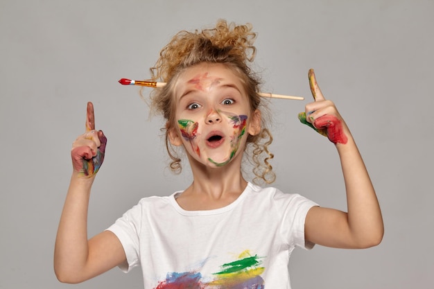 Beautiful little girl having a brush in her chic curly blond hair, wearing in a white t-shirt, with a painted fingers, is acting like she has got an idea, on a gray background.