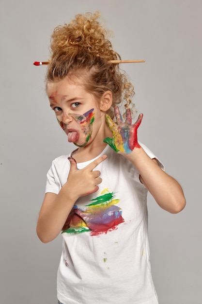 Beautiful little girl having a brush in her lovely haircut, wearing in a white smeared t-shirt. She is posing with a painted hands and cheeks, pointing on her hand and showing a tongue, on a gray back