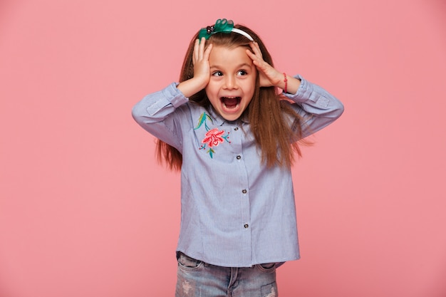 Beautiful little girl reacting emotionally grabbing head with both hands being delighted and shocked