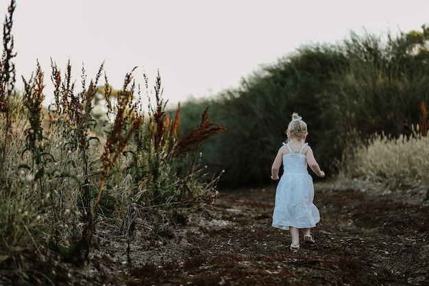 Beautiful little girl in a white dress walking along a country road at sunset