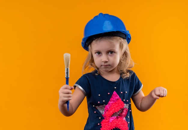 Una bella bambina con capelli biondi che indossa camicia blu navy e casco blu che tiene il pennello blu su una parete arancione