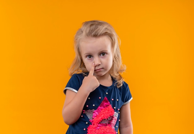 A beautiful little girl with blonde hair wearing navy blue shirt pointing at her face with index finger on an orange wall