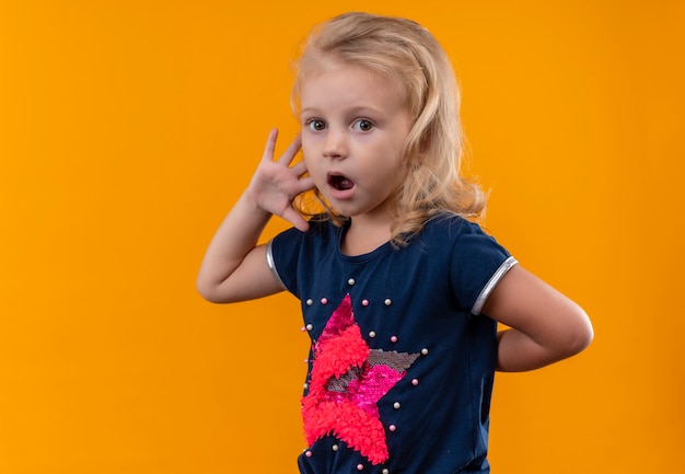 Foto gratuita una bella bambina con i capelli biondi che indossa la camicia blu navy che mostra un'espressione sorpresa mentre osserva su una parete arancione