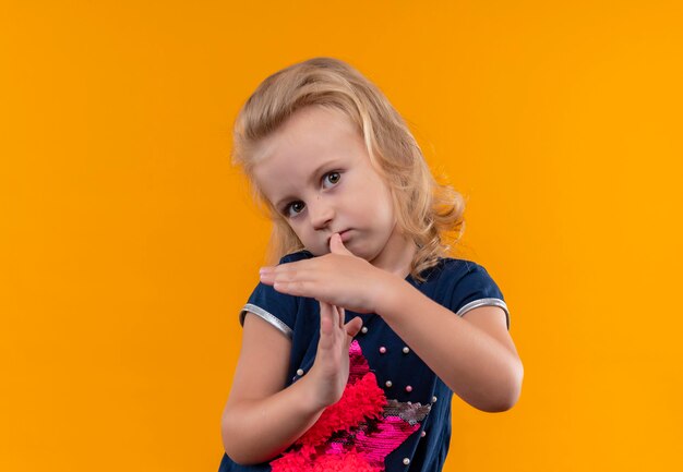 A beautiful little girl with blonde hair wearing navy blue shirt showing timeout sign with fingers on an orange wall
