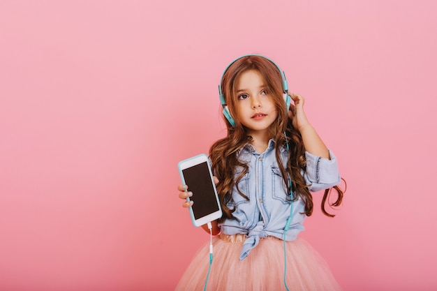 Beautiful little girl with long brunette hair with phone listening to music through blue headphones isolated on pink background. Cheerful mood of young child, enjoying music