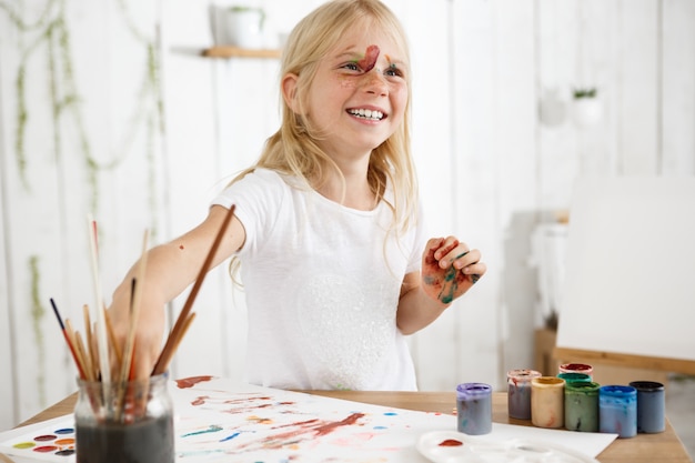 Beautiful little girl with straight blonde hair, freckles and paint on her face, laughing and having fun. Kids' art activities.