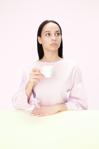 ピンクのスタジオに座って、コーヒーを手に持って悲しそうに見える美しい孤独な女性。