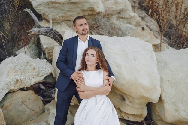 красивая длинношерстная невеста в белом платье с мужем на пляже возле больших камней