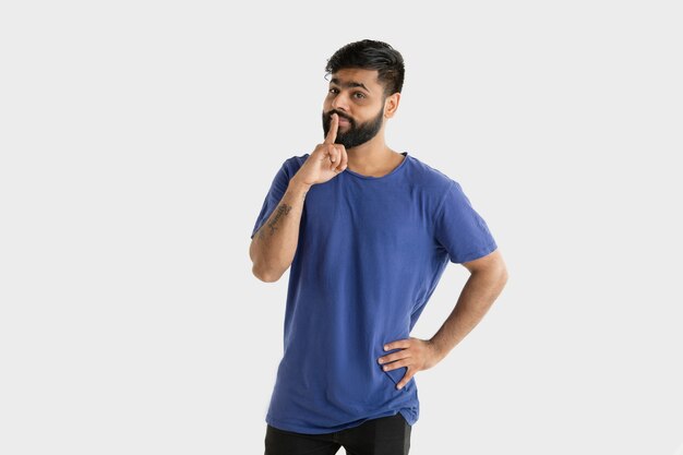 白いスタジオの背景に分離された美しい男性の半分の長さの肖像画。青いシャツを着た若い感情的なヒンドゥー教の男。顔の表情、人間の感情、広告のコンセプト。考えて、秘密をささやきます。