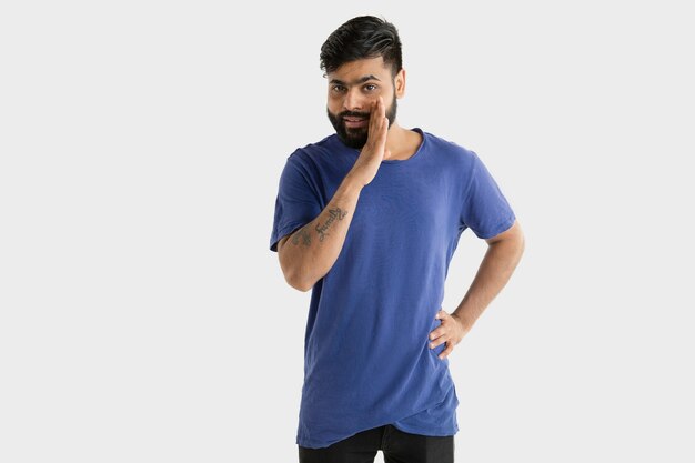 Beautiful male half-length portrait isolated on white wall. Young emotional hindu man in blue shirt. Facial expression, human emotions, advertising concept. Smiling, whispering a secret.