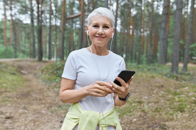 Beautiful middle aged Caucasian woman with short hair enjoying summer morning outdoors, going to have cardio workout, choosing music tracks on mobile phone