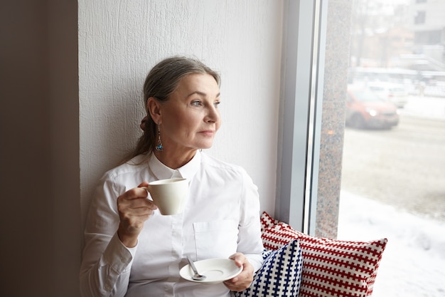 Beautiful middle aged female with gray hair and blue eyes sitting at cafeteria on windowsill, enjoying morning coffee, holding cup and looking through window, havign thoughful facial expression