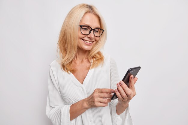 Beautiful middle aged woman chats on mobile phone glad to receive message from daughter smiles happily wears neat white blouse and eyeglasses