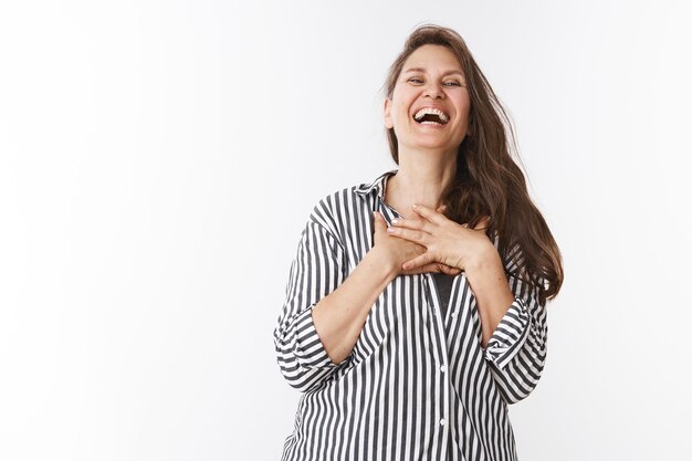 Beautiful middle aged woman in striped trendy blouse chuckling, laughing out loud holding hands on chest from amazement shaking from laugh and joy bending head backwards amused over white wall