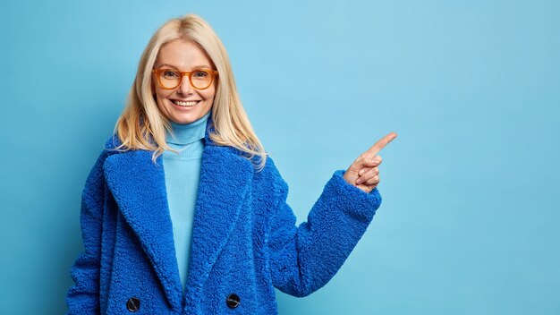 Beautiful middle aged woman with blonde hair wears spectacles and warm blue coat pointing at copy space