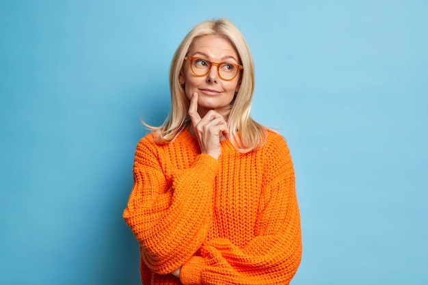 Beautiful middle aged wrinkled woman keeps index finger on cheek stands in thoughtful pose daydreams about something wears knitted sweater.