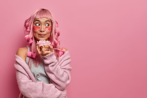 Beautiful mixed race woman has pink hair choppy bangs, wears beauty pads to reduce wrinkles, treats herself with delicious glazed doughnut, makes hairstyle