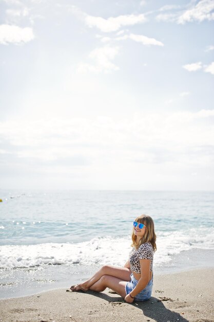 ジーンズの短いヒョウのシャツとサングラスを身に着けている海のビーチでリラックスした美しいモデル