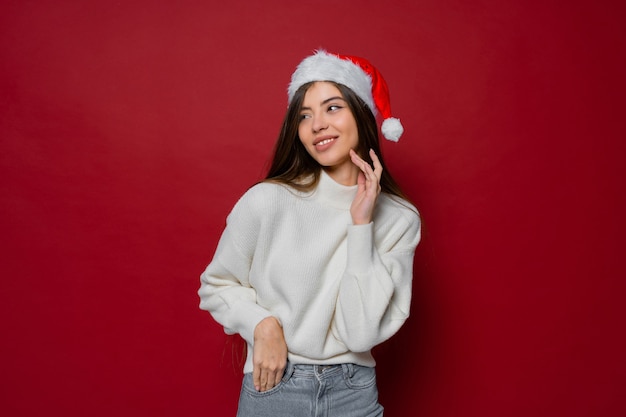 Beautiful model in santa hat and white cozy sweater posing on red