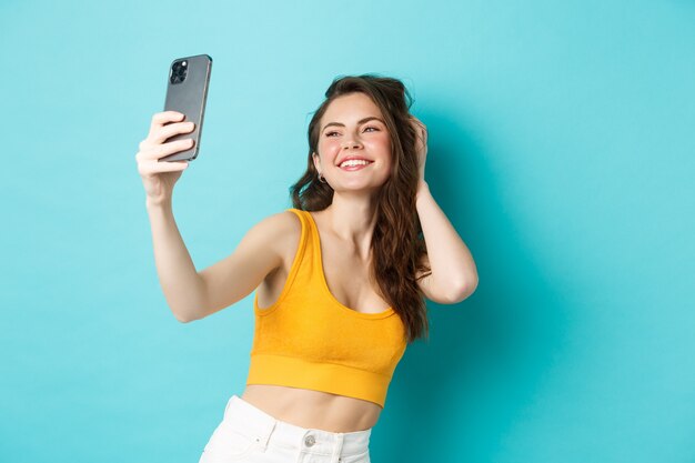 Beautiful modern girl taking selfies on vacation, posing in summer clothes and looking at smartphone camera, standing against blue background.