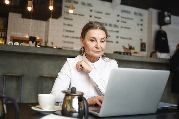 아름 다운 현대 중년 유럽 여성 프리랜서 휴대용 컴퓨터에서 멀리 일하고, 카페테리아에 앉아 카푸치노를 마시고. 카페에서 원격 작업을 위해 노트북을 사용하는 노인 여성 작가
