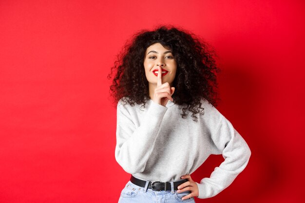 Beautiful modern woman with curly hair and makeup, shushing and smiling, telling a secret, making surprise and smiling, standing over red background