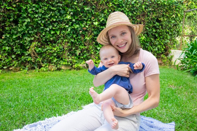 Beautiful mom in hat sitting on plaid with newborn in park, smiling and holding him. Little baby laughing and waving legs and hands. Summer family time, sunny days and fresh air concept