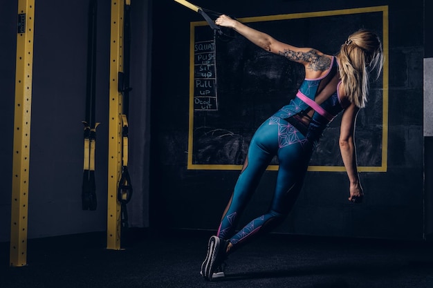 ジムでTRXシステムで運動をしている彼女の腕に入れ墨をしたスポーツウェアの美しい筋肉のブロンドの女性。 TRXのコンセプト。
