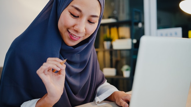 Beautiful muslim lady in headscarf casual wear using laptop in living room at night house.