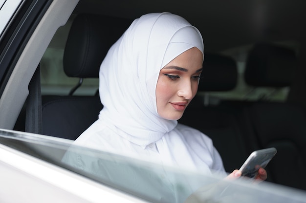 Красивая мусульманская женщина в белом хиджабе сидит на заднем сиденье автомобиля и использует смартфон