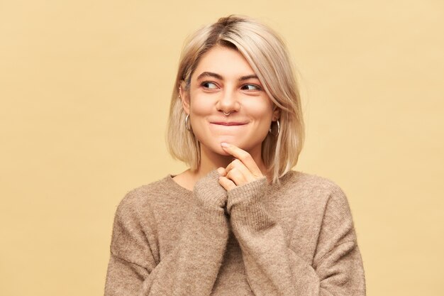 Beautiful mysterious European girl in stylish cozy sweater having amazed enigmatic facial expression, looking away and smiling, coming up with idea, thinking about plan isolated at blank  wall