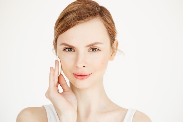 Beautiful natural blonde girl cleaning face with cotton sponge smiling . Cosmetology and spa.