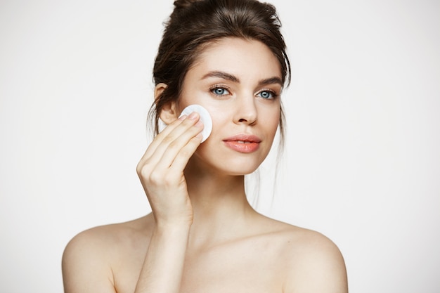 Beautiful natural brunette girl cleaning face with cotton sponge smiling over white background. cosmetology and spa.